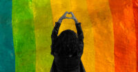 A person holds up a heart sign against a rainbow background.