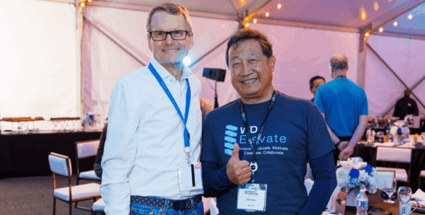 Two WD employees celebrating together at WD Elevate event in tent setting, one wearing WD Elevate branded shirt and giving thumbs up gesture.